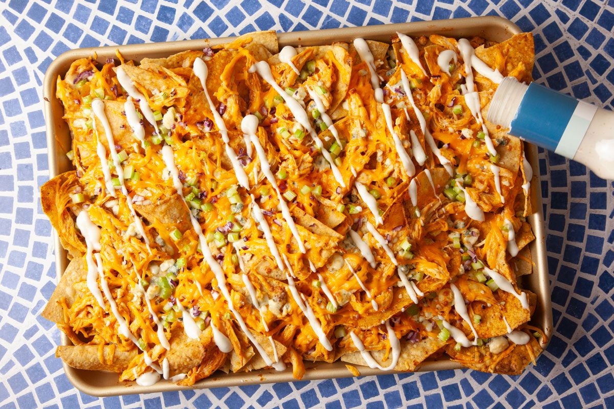 Overhead action shot of ranch or blue cheese dressing being drizzled over fully assembled Buffalo Chicken Nachos. The creamy drizzle adds contrast and enhances the bold Buffalo flavor profile.