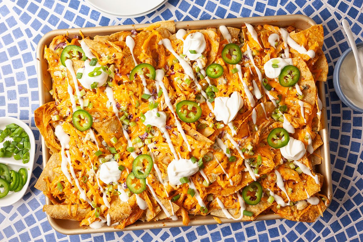 Overhead hero image of fully garnished Buffalo Chicken Nachos topped with sliced jalapeños, dollops of sour cream, chopped green onions, celery, and red onion. A vibrant and flavorful appetizer perfect for sharing.