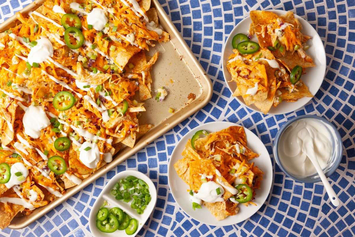 Overhead entertaining scene featuring Buffalo Chicken Nachos served on a sheet pan with individual plated portions and small bowls of dressing and jalapeños. Captures the finished recipe ready for sharing at gatherings.