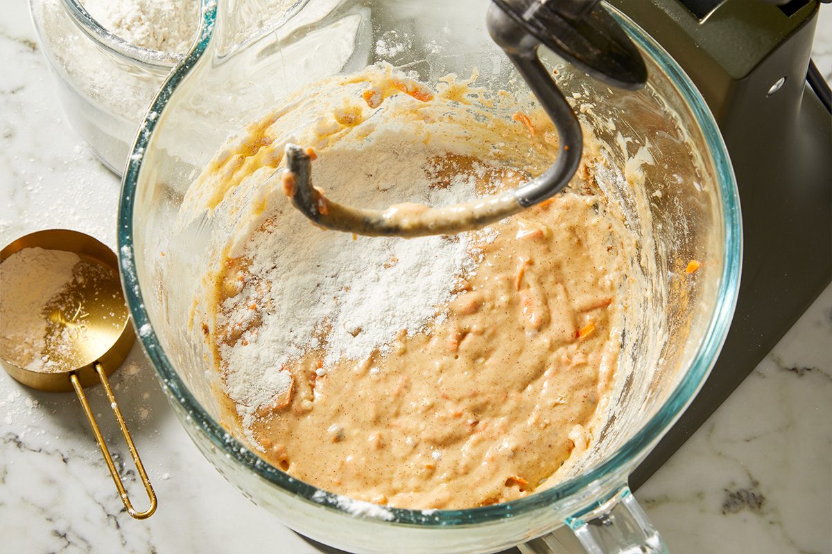 Overhead view of carrot cake cinnamon roll dough ingredients being mixed in a glass bowl with a dough hook, showing shredded carrots incorporated into the dough.