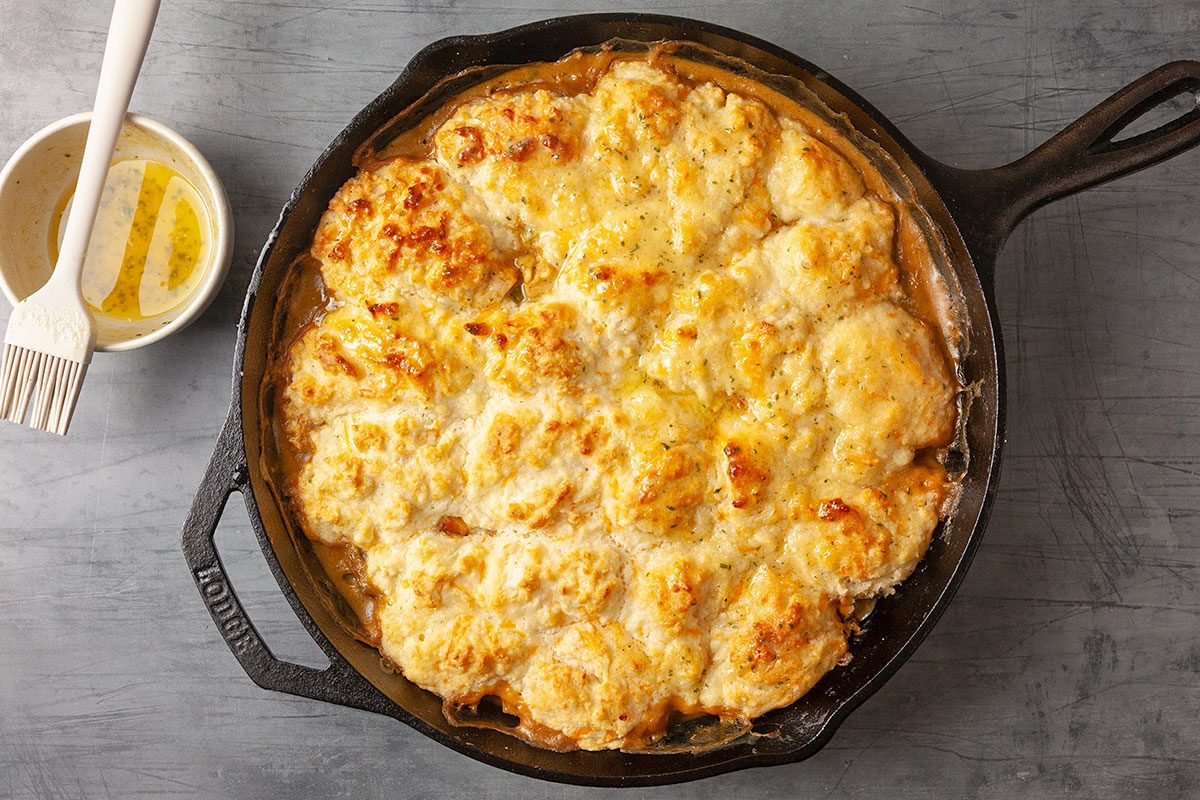 Overhead horizontal image of a freshly baked Cheddar Bay Chicken Pot Pie with a golden biscuit topping, shown with a pastry brush nearby, suggesting a butter or herb finish.