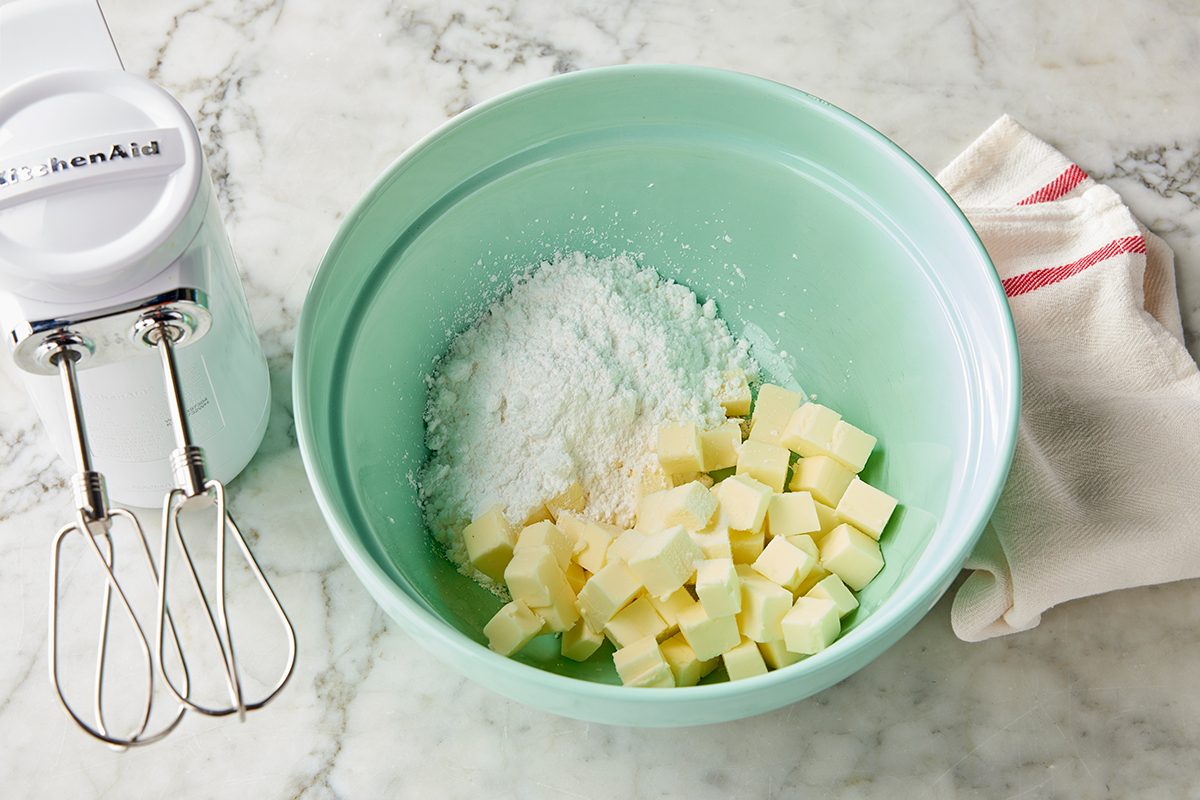 A light green mixing bowl with cubed butter and powdered sugar inside, next to a white electric hand mixer and a folded kitchen towel on a marble countertop.