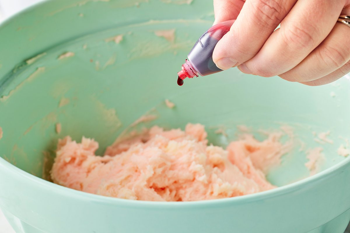 A hand squeezes red food coloring from a bottle into a light green bowl containing a pale dough or batter mixture.