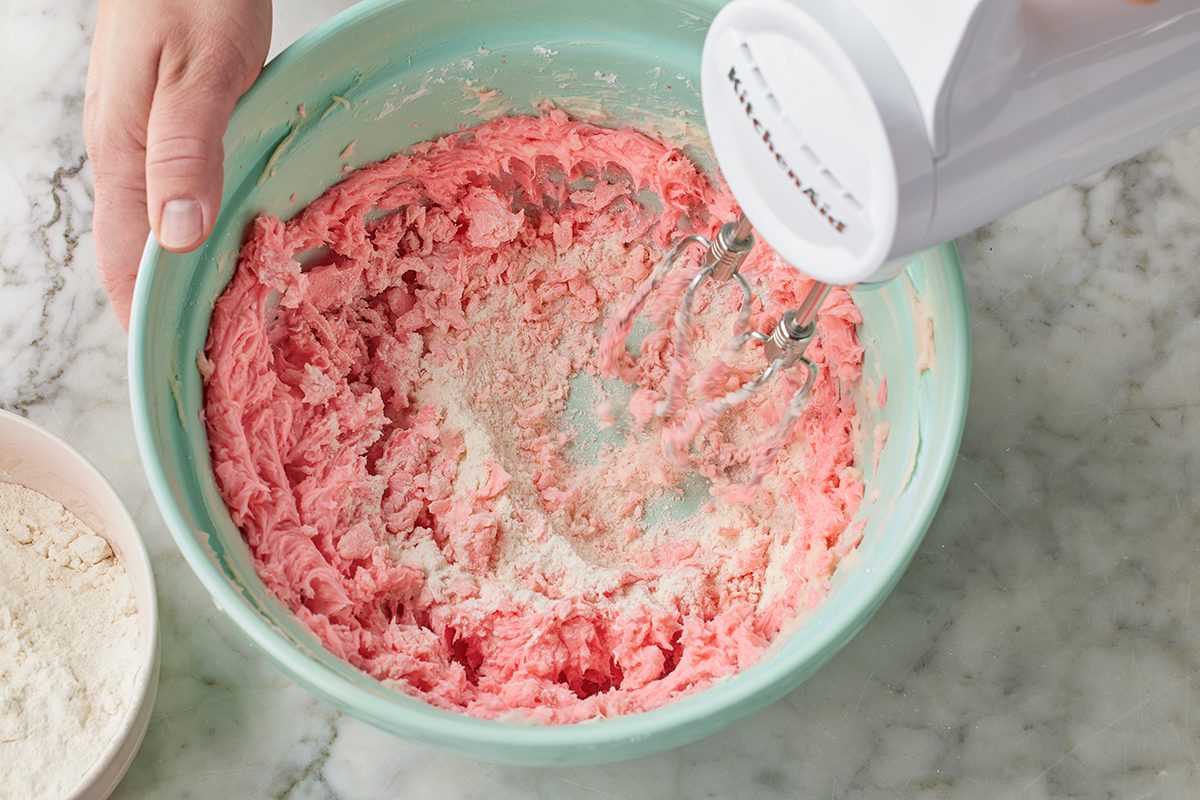 A person uses a hand mixer to blend pink batter in a light blue mixing bowl on a marble countertop, with a bowl of flour nearby.