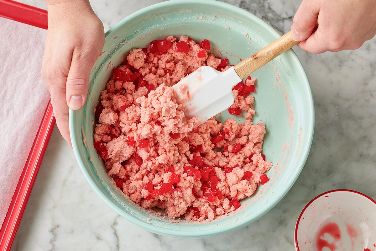 A person uses a spatula to mix a pink, crumbly dough with red chunks in a green bowl on a marble countertop.