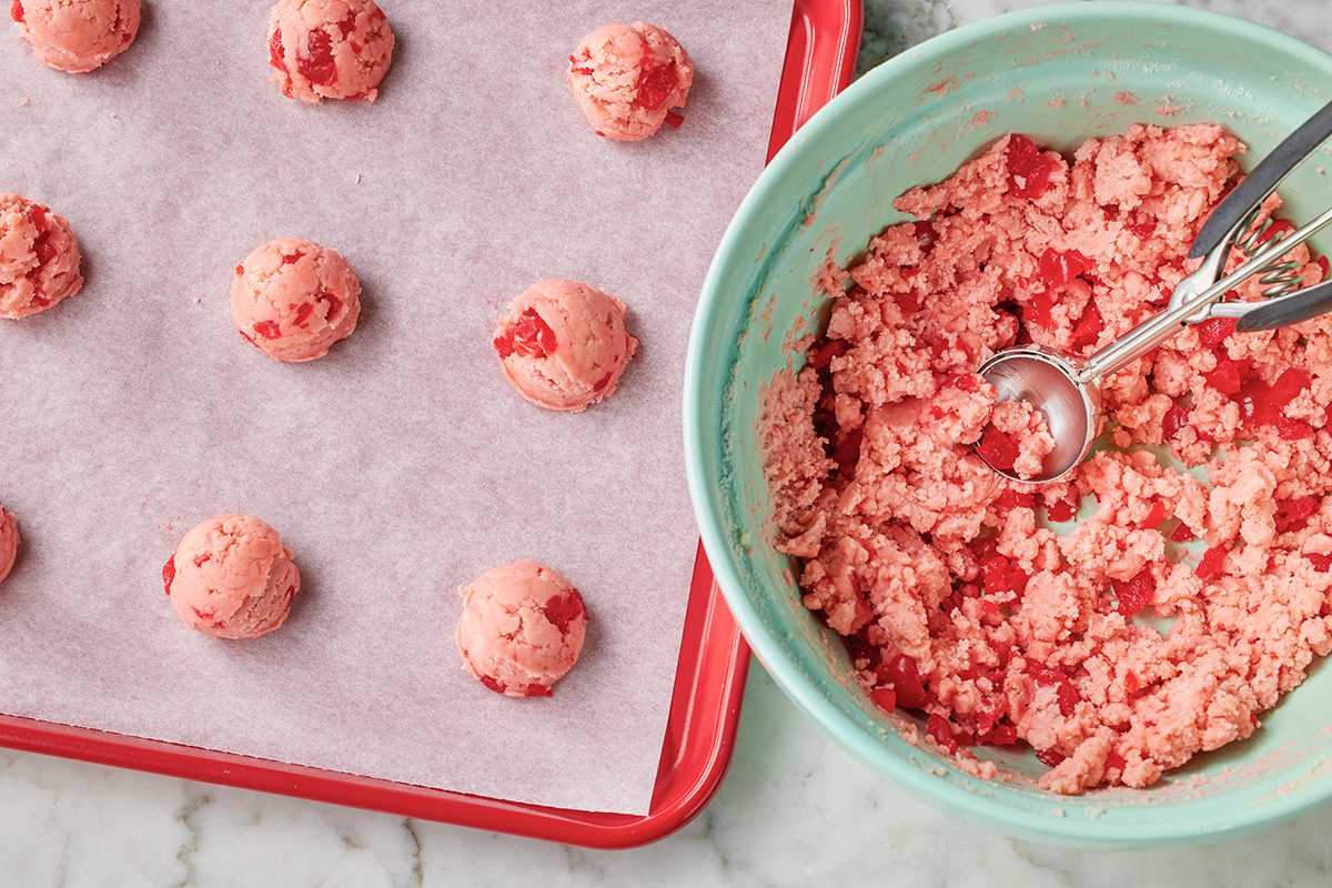 A baking sheet lined with parchment paper holds rows of pink cookie dough balls. Beside it, a green mixing bowl contains more pink dough with a metal scoop resting inside.