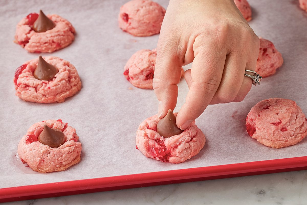 A hand presses a chocolate kiss into the center of a pink cookie on a baking sheet lined with parchment paper, surrounded by similar cookies.