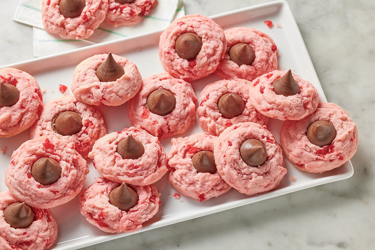 A white tray holds several pink cookies, each topped with a chocolate candy drop in the center. Some cookies have no chocolate topping. The surface beneath the tray is marble.