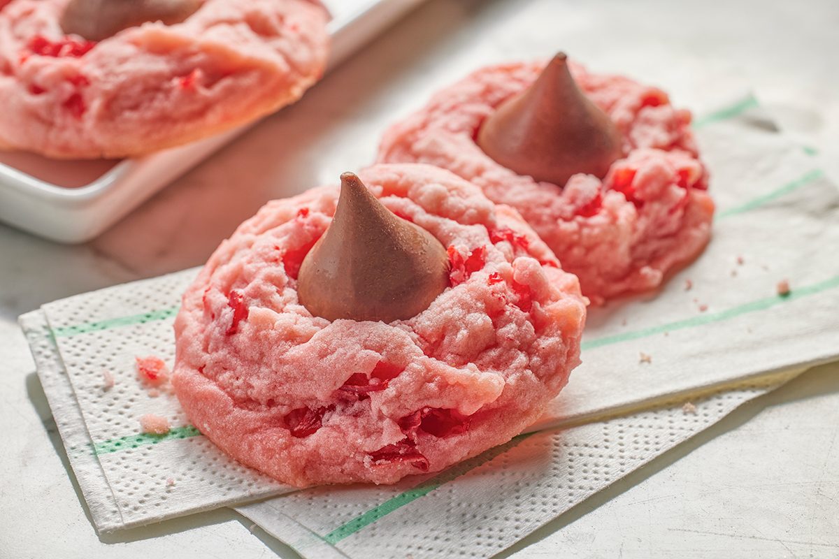 Two pink cherry cookies topped with chocolate candy kisses rest on a white napkin, with a tray of more cookies in the background.
