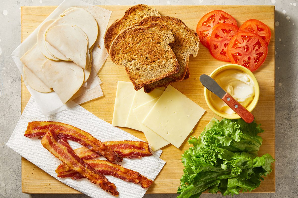 A wooden board with sliced turkey, toasted bread, cheese slices, tomato slices, lettuce, crispy bacon on a paper towel, and a small bowl of mayonnaise with a spreading knife.