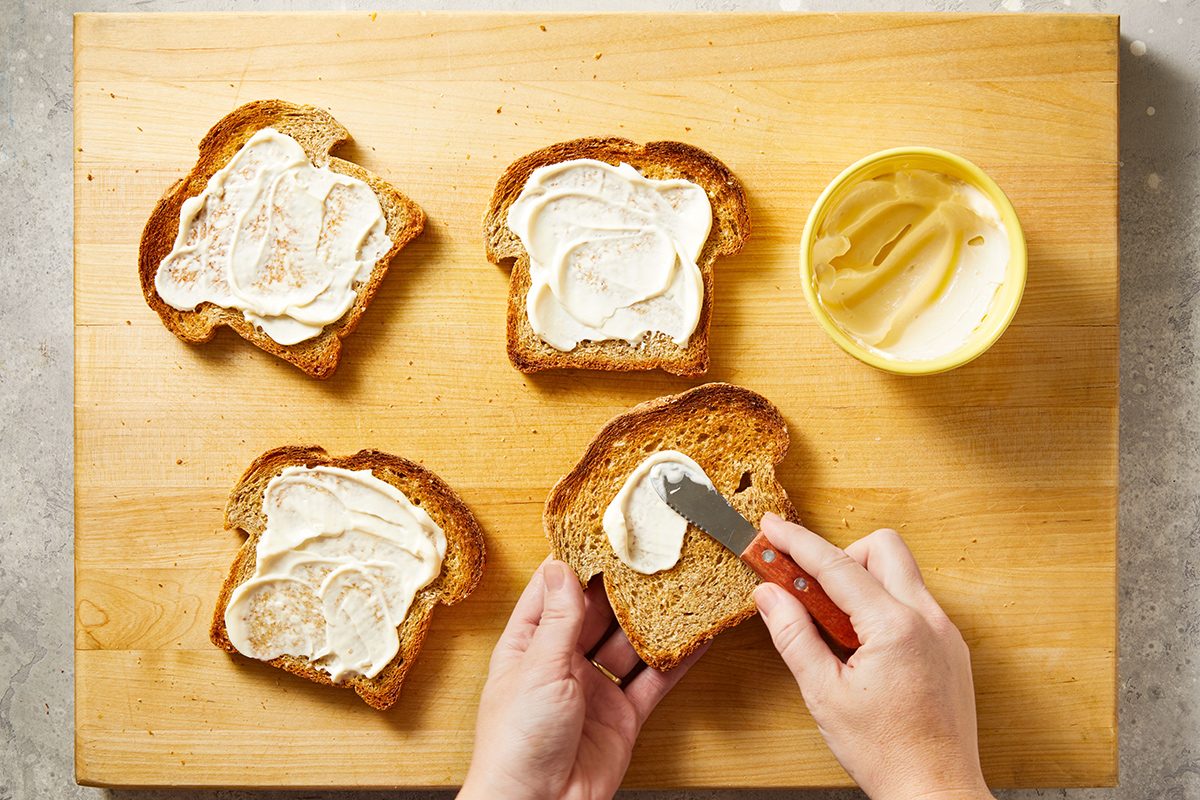 Four slices of bread on a wooden board, each being spread with a creamy spread. A person spreads more on one slice with a butter knife, and an open container of spread is nearby.
