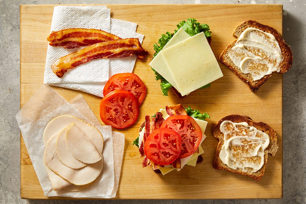 Sandwich ingredients on a wooden board: cooked bacon strips, sliced tomatoes, lettuce, cheese slices, deli meat, and two slices of bread spread with mayonnaise, one topped with sandwich fillings.