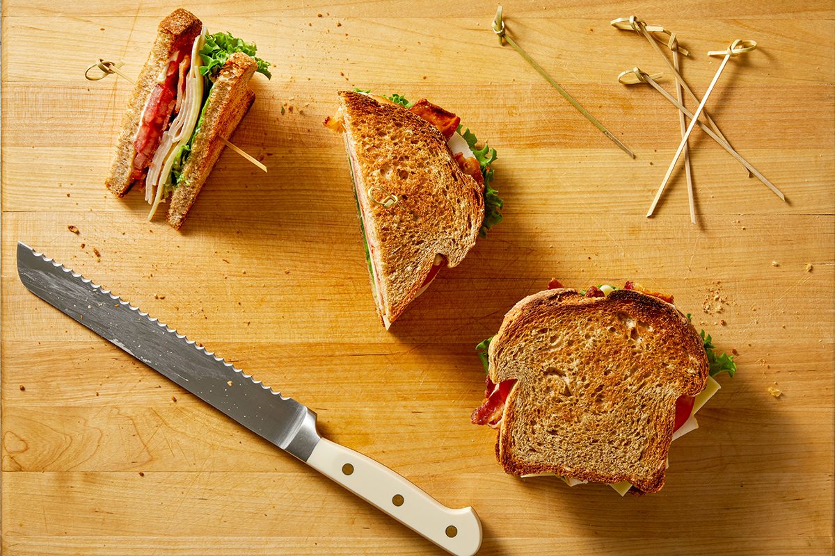 A wooden cutting board with a bread knife, two sandwiches—one whole and one cut in half—and a few wooden sandwich picks scattered nearby.