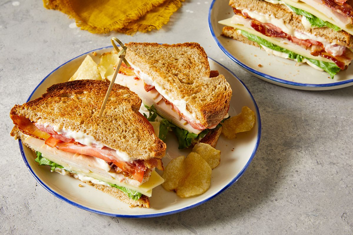 Two plates with club sandwiches made of toasted bread, lettuce, tomato, bacon, turkey, and cheese, served with potato chips on a light gray surface. One sandwich is held together with a toothpick.