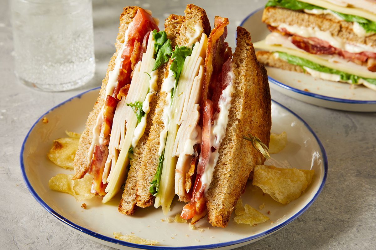 A club sandwich stacked with turkey, bacon, lettuce, tomato, and cheese on toasted bread, served on a plate with potato chips. A glass of water and another sandwich are in the background.