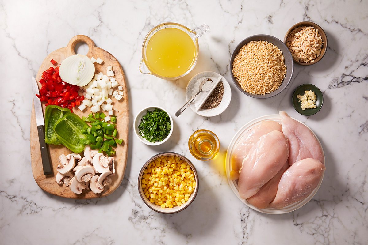 Overhead shot of a marble countertop with raw chicken breasts, chopped vegetables, corn, chicken broth, herbs, oil, rice, seasonings, and a knife on a wooden cutting board, ready for meal preparation;