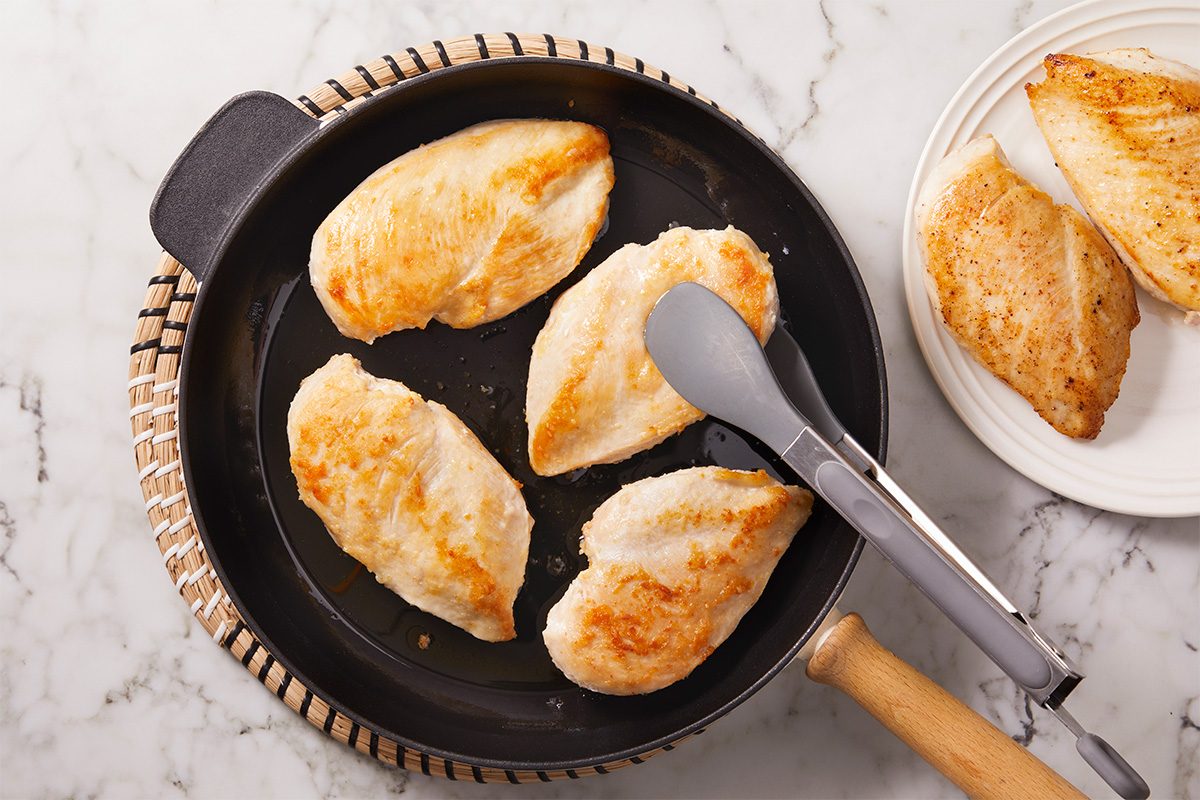 Overhead shot of four cooked chicken breasts in a black frying pan with metal tongs, with a plate of additional chicken on the side on a white marble surface;
