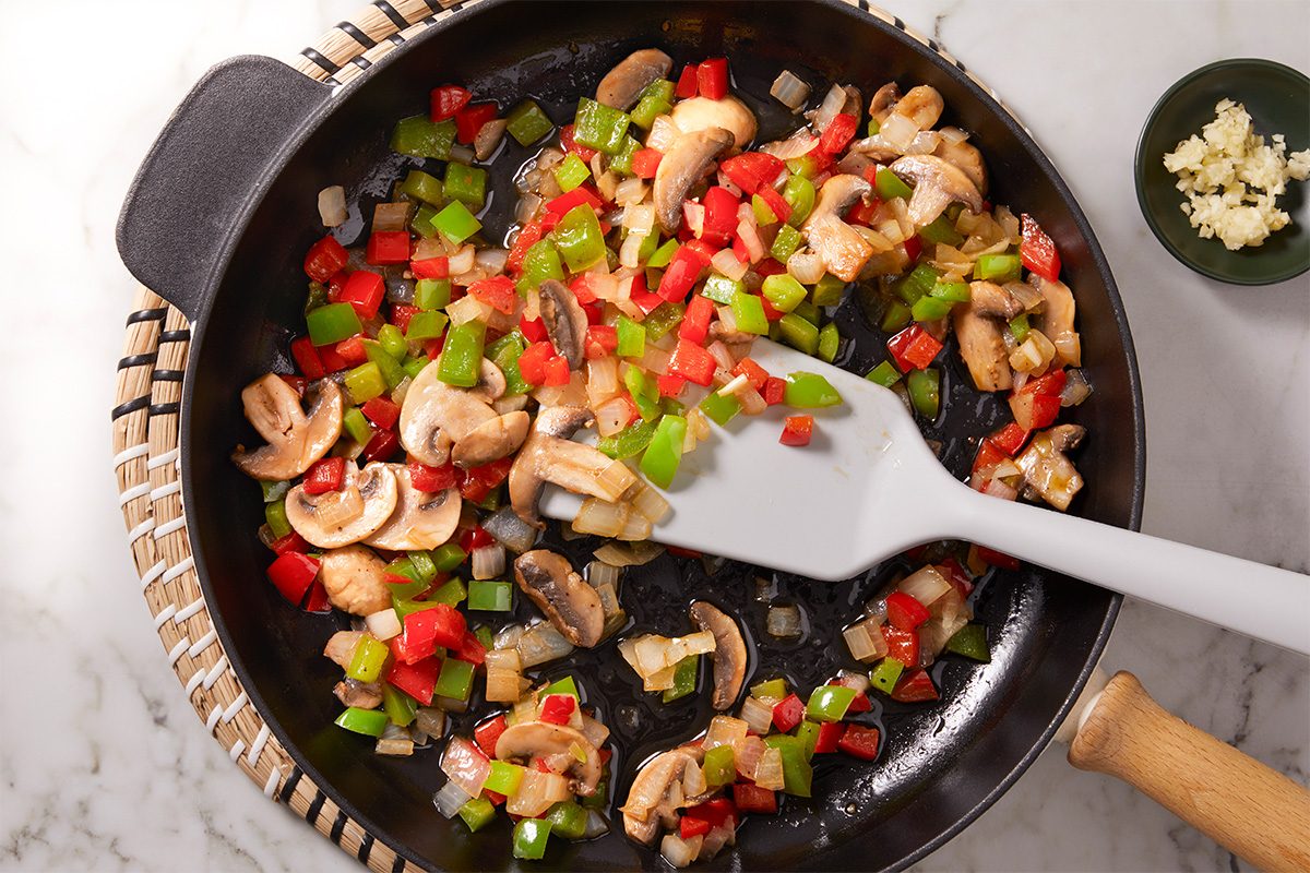 Overhead shot of a skillet filled with sautéed mushrooms, diced red and green bell peppers, and onions being stirred with a white spatula, with a small bowl of chopped garlic nearby;