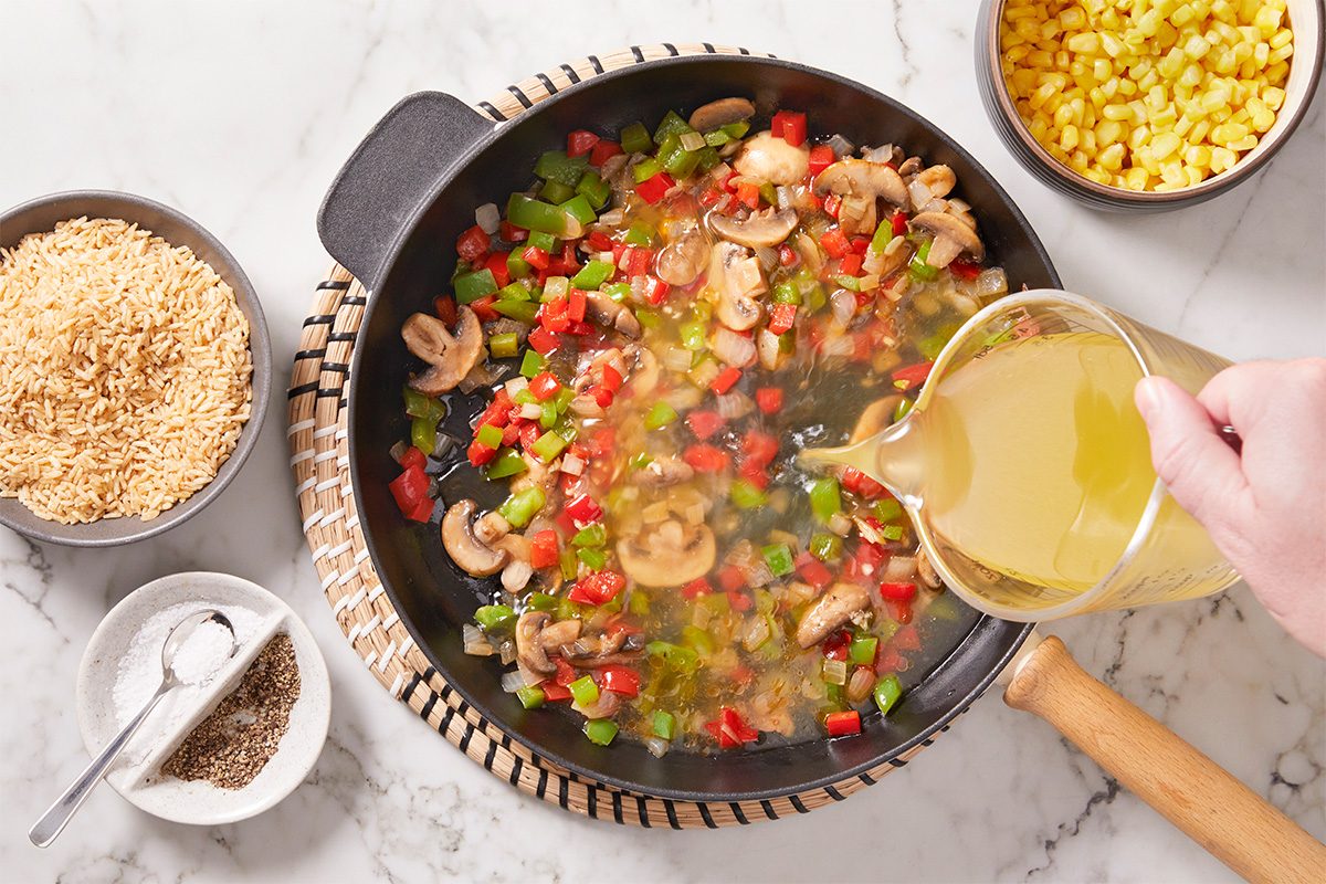 Overhead shot of a hand pouring broth into a skillet with chopped red and green bell peppers, mushrooms, and onions, with bowls of uncooked rice, corn, and seasonings nearby on a marble countertop;