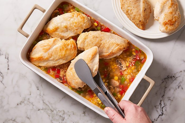 Overhead shot of a hand using tongs to place cooked chicken breasts into a baking dish filled with rice, vegetables, and broth, with additional chicken on a plate nearby on a marble countertop;