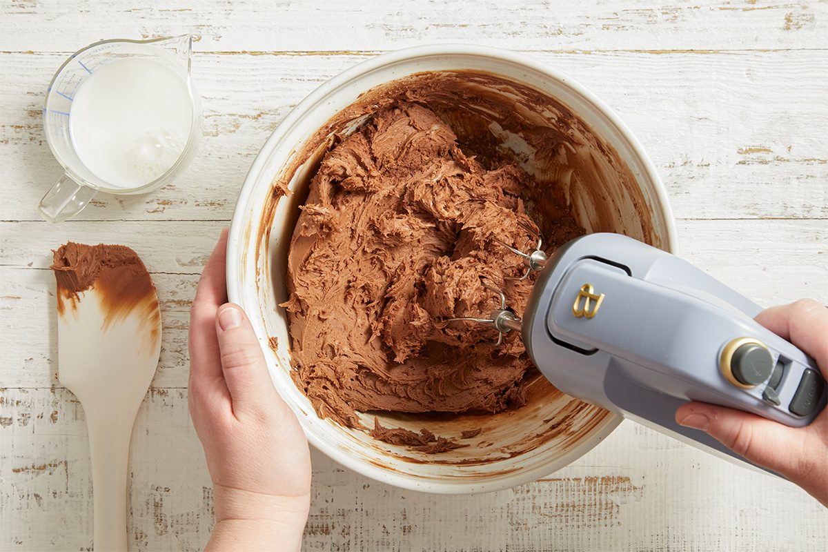 Overhead action shot of chocolate cake batter being blended with a hand mixer in a large bowl. The mixture appears thick and smooth as ingredients are fully incorporated.