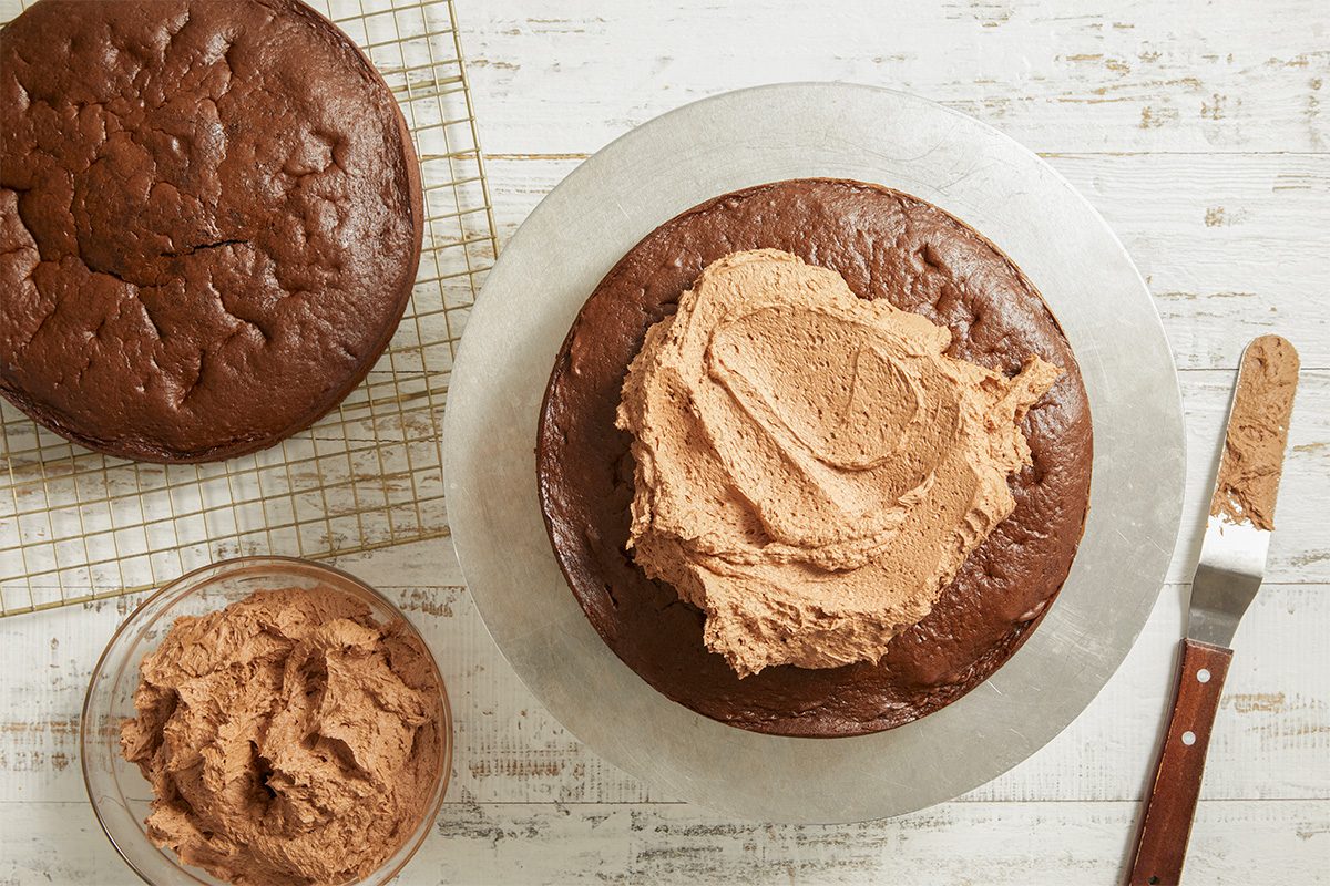 Overhead image of chocolate frosting being spread onto a cooled cake layer. A spatula smooths the creamy frosting evenly across the surface.