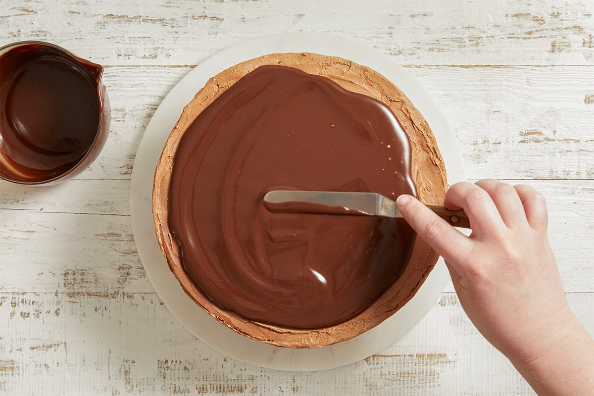 Overhead view of glossy chocolate ganache being spread over the top of a frosted cake, creating a smooth, rich finish.