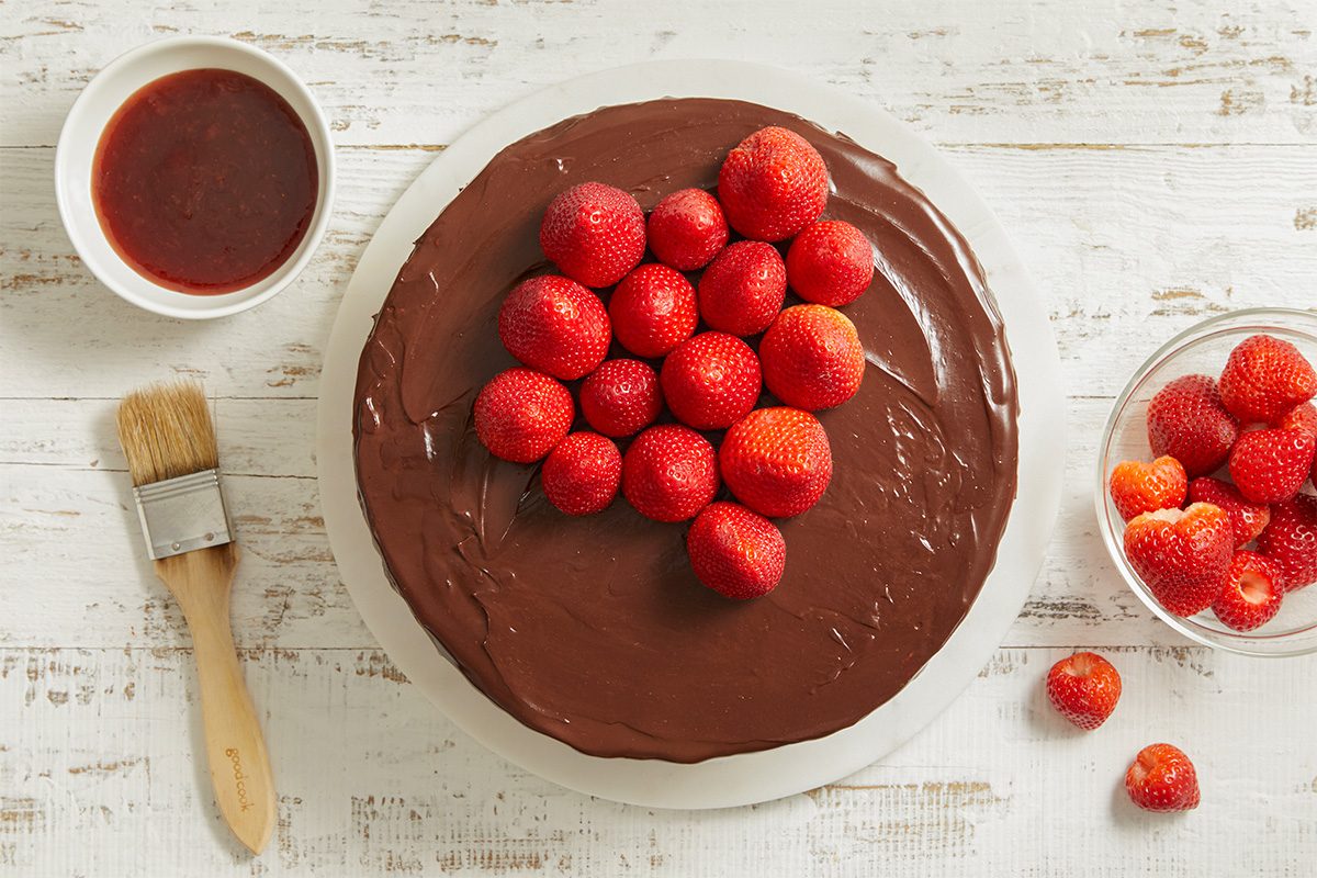 Overhead view of fresh strawberries arranged on top of the chocolate-glazed cake. A small bowl of strawberry jam sits nearby for optional glazing.