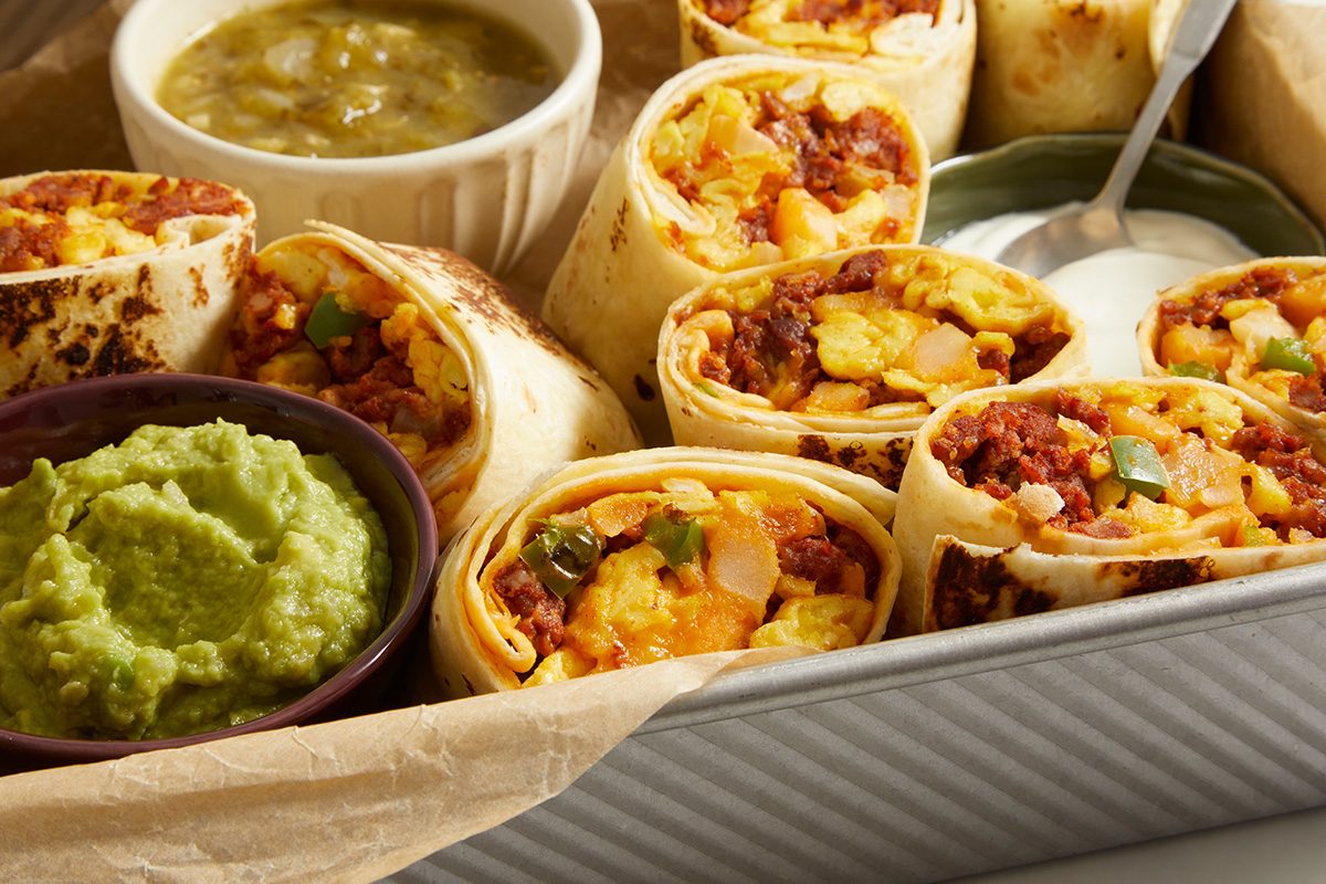 A tray of breakfast burritos filled with eggs, sausage, and vegetables, served with sides of guacamole, salsa, and sour cream in small bowls.
