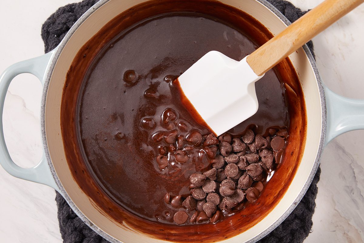A white pot filled with smooth, melted chocolate and chocolate chips being stirred with a white spatula. The pot is resting on a dark knitted trivet.