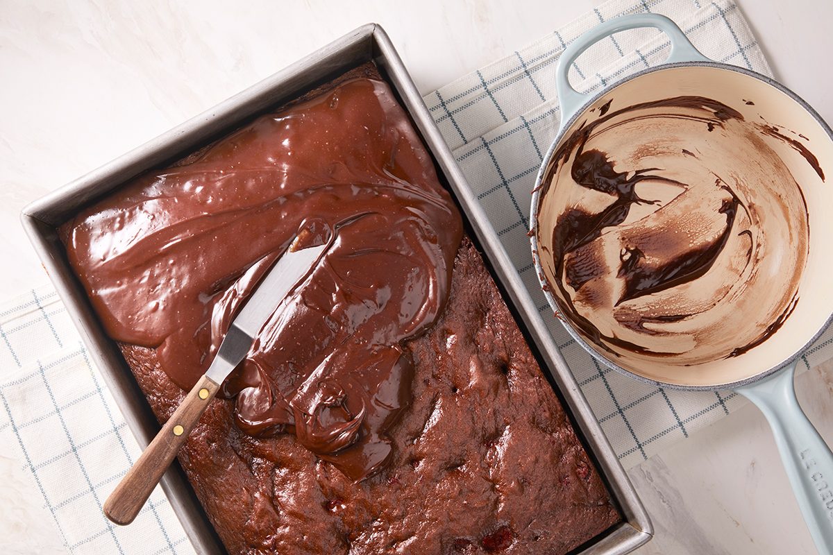 A baking pan of chocolate cake is being frosted with chocolate icing using a spatula. Next to it is a mixing bowl with leftover chocolate frosting and a wire cooling rack underneath.