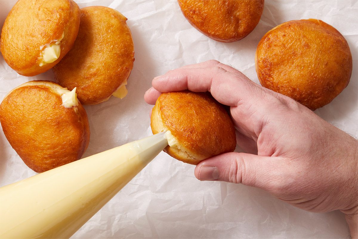 A horizontal overhead image showing fried doughnuts being filled with pastry cream using a piping bag. The image demonstrates filling Crème Brûlée Doughnuts with rich custard.