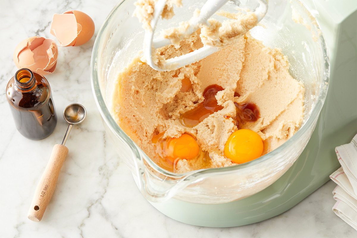 In a large bowl, beat butter and sugars until blended. Beat in eggs and vanilla. Overhead step-by-step image of softened butter, brown sugar and granulated sugar creamed together in a glass mixing bowl. Eggs and vanilla extract are added to the mixture on a light marble surface, with baking tools nearby. This image highlights the beginning stage of making Easter Chocolate Chip Cookies.