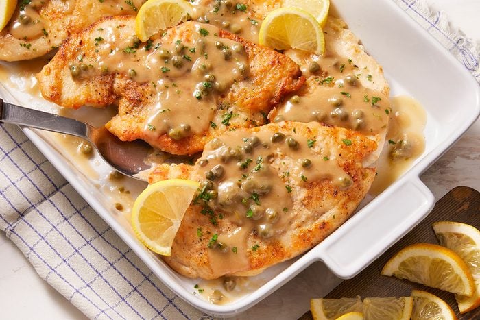 Golden chicken breasts topped with a creamy caper sauce, garnished with chopped herbs and lemon slices, served in a white baking dish with a serving spoon on the side.