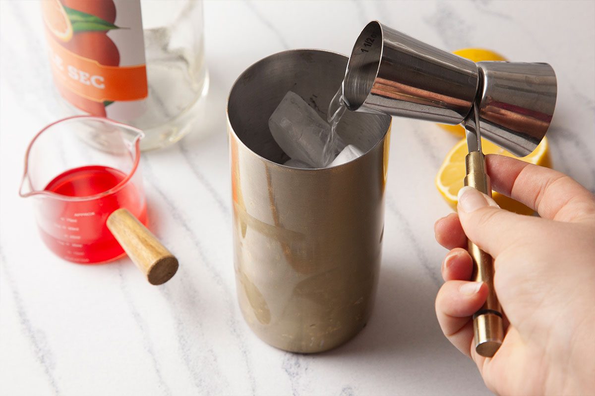 Overhead step-by-step preparation image of a Gin Daisy cocktail showing gin being measured with a metal jigger and poured over ice into a stainless steel cocktail shaker. Fresh lemon halves, a small measuring cup of grenadine syrup, and bar tools rest on a light marble surface, illustrating the beginning of the cocktail mixing process.
