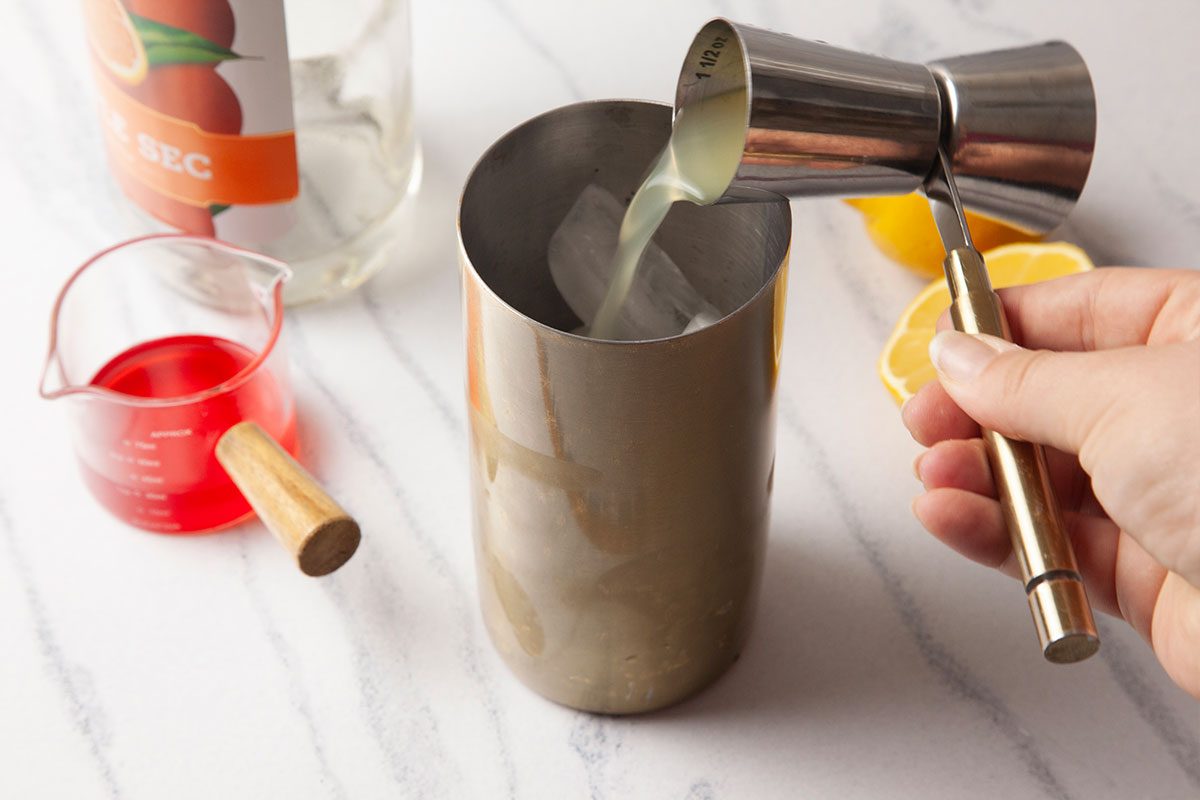 Overhead process image of lemon juice and orange liqueur being measured and poured into a cocktail shaker filled with ice for a Gin Daisy. The scene includes a jigger, fresh lemon halves, and bar tools arranged on a marble surface, capturing the layered building of the cocktail.