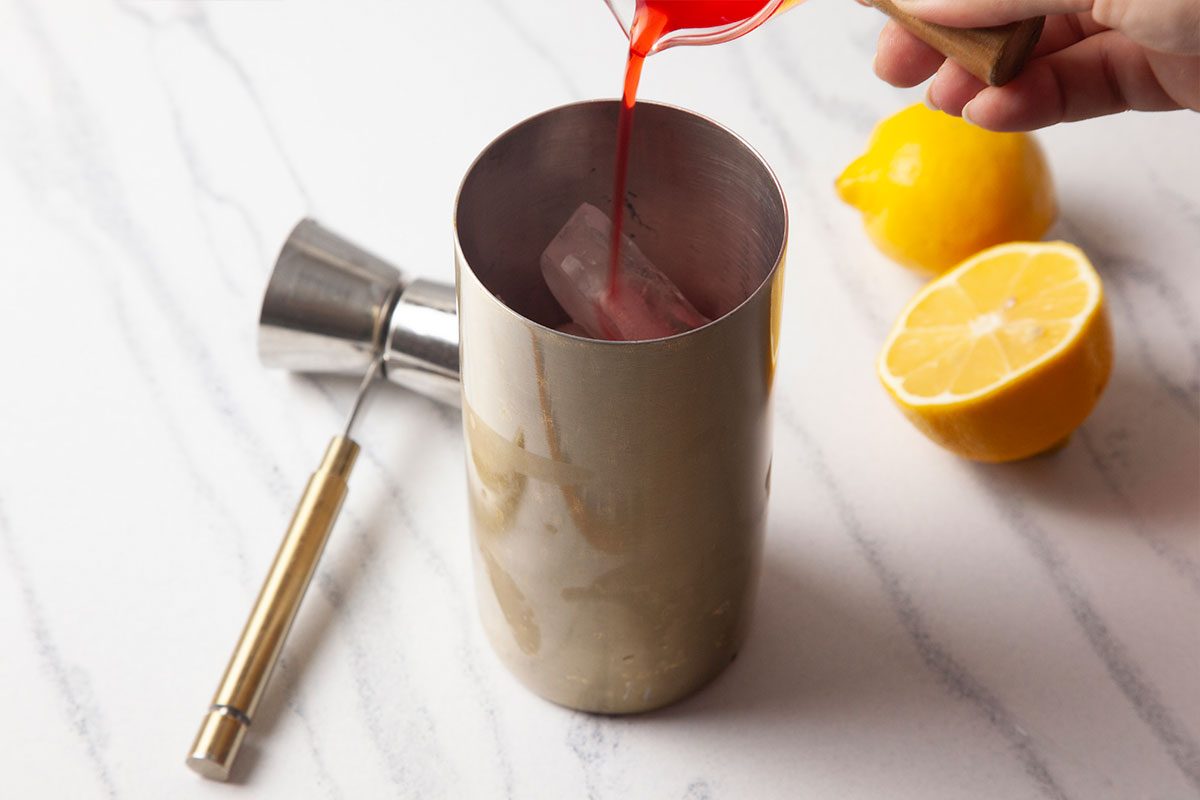 Overhead step-by-step image of bright red grenadine syrup being poured into a cocktail shaker containing gin, citrus, and ice. Fresh lemons and bar tools surround the shaker on a marble countertop, highlighting the vibrant color addition that defines a Gin Daisy.