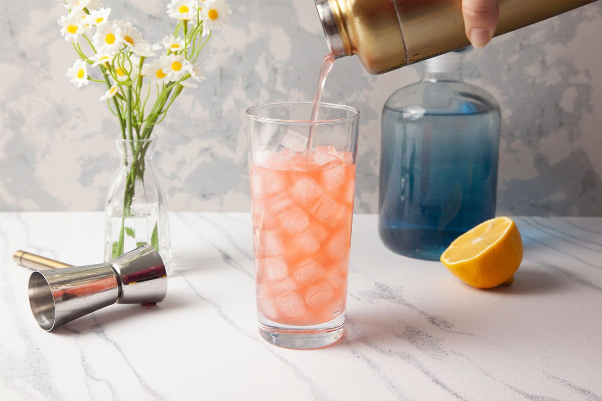 Overhead action image of a pink Gin Daisy being strained into a tall glass filled with ice and topped with club soda. A blue glass bottle, lemon halves, and fresh flowers add styling elements on a marble background, emphasizing the refreshing finish.