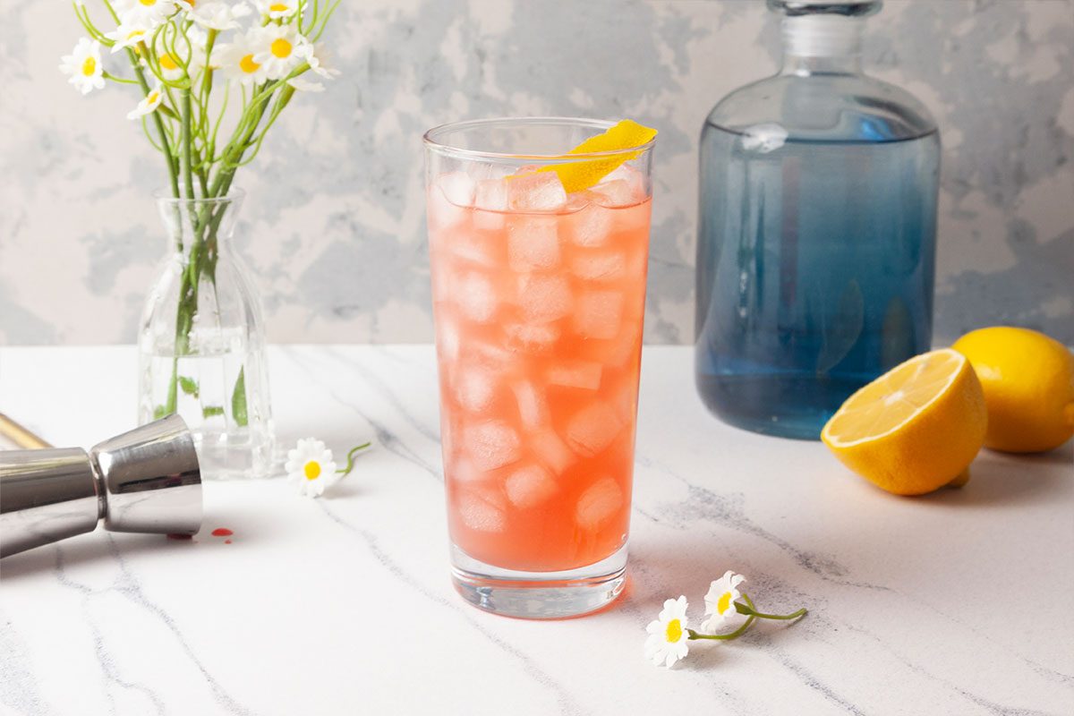 Overhead styled image of a finished Gin Daisy cocktail served in a tall glass filled with ice and garnished with a lemon peel twist. A blue bottle, lemon halves, chamomile flowers, and bar tools complete the scene on a marble surface.