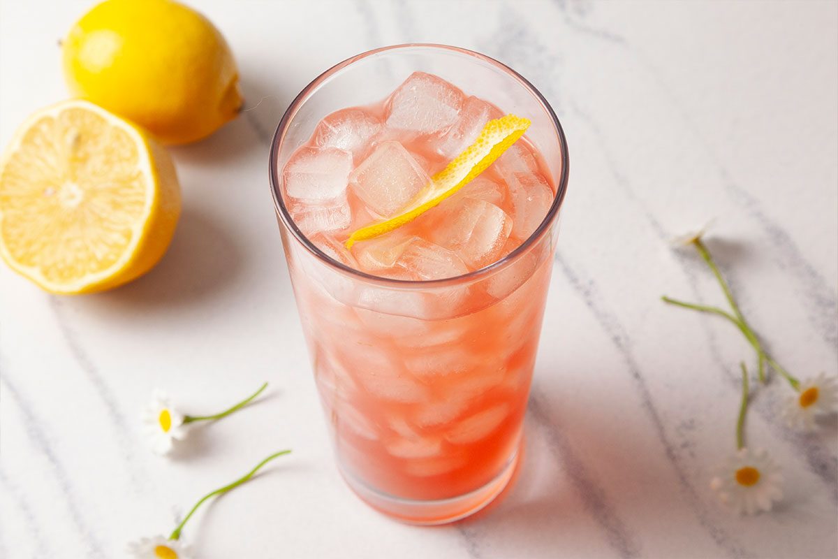 Push-and-pulls styled overhead close-up of a Gin Daisy cocktail highlighting the pink hue, ice cubes, and lemon peel garnish. Fresh lemon halves and delicate flowers surround the glass, emphasizing texture and color.