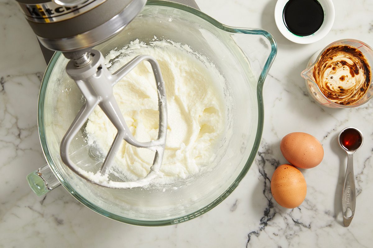 A stand mixer with creamed ingredients in a glass bowl sits on a marble countertop, surrounded by two eggs, a cup of espresso, a bowl of liquid, and a small dish of vanilla extract.