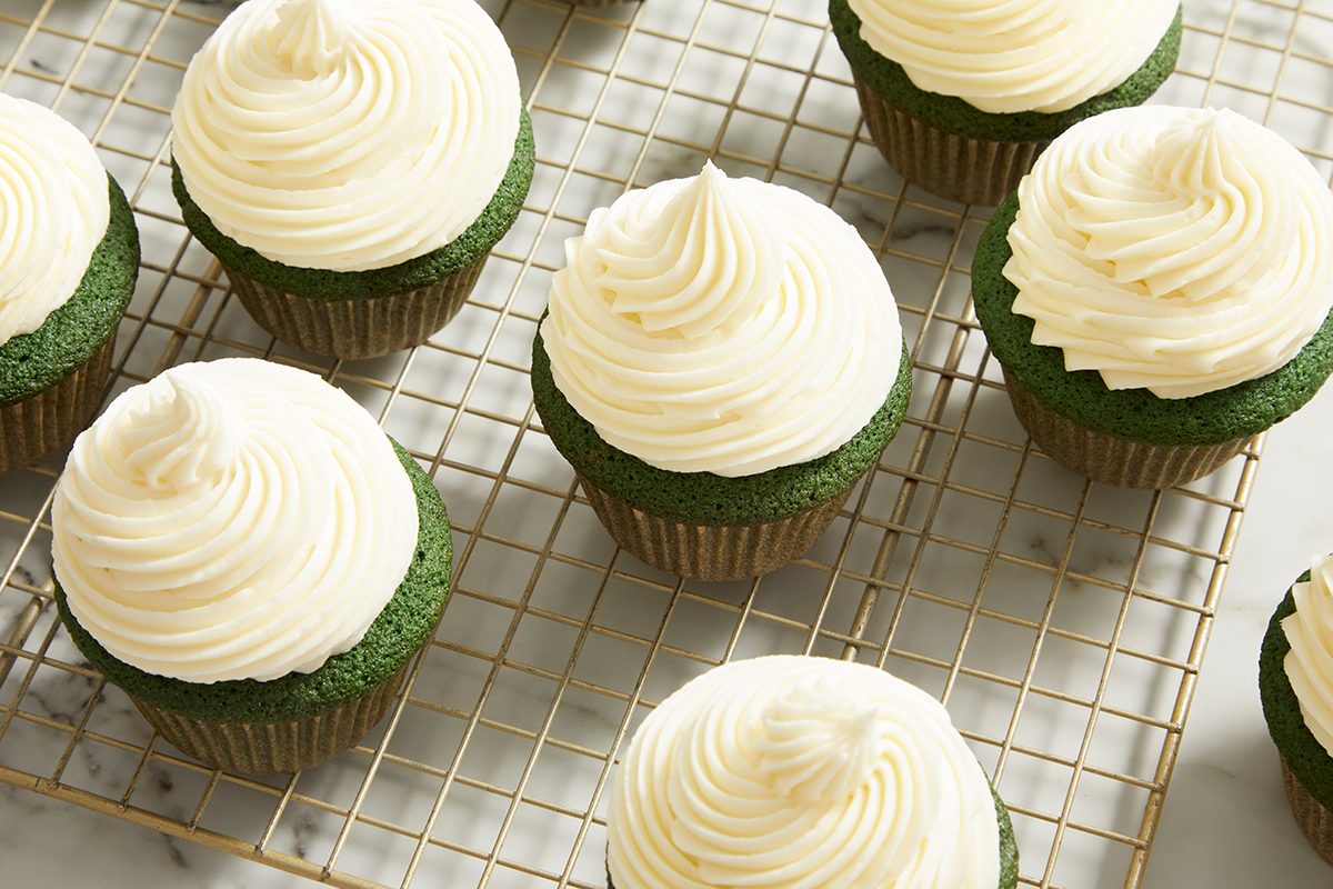 Green cupcakes with swirled white frosting are arranged on a cooling rack. The cupcakes are evenly spaced and the rack sits on a marble surface.