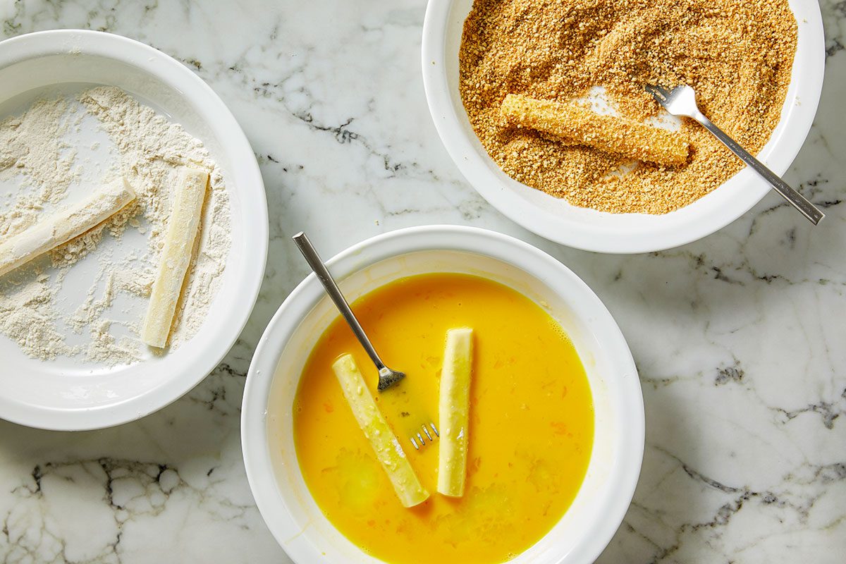 Overhead shot of three bowls on a marble surface; one filled with flour, one with beaten eggs, and one with breadcrumbs; cheese sticks are being coated with one placed in each bowl; forks rest in the bowls for dipping and coating;