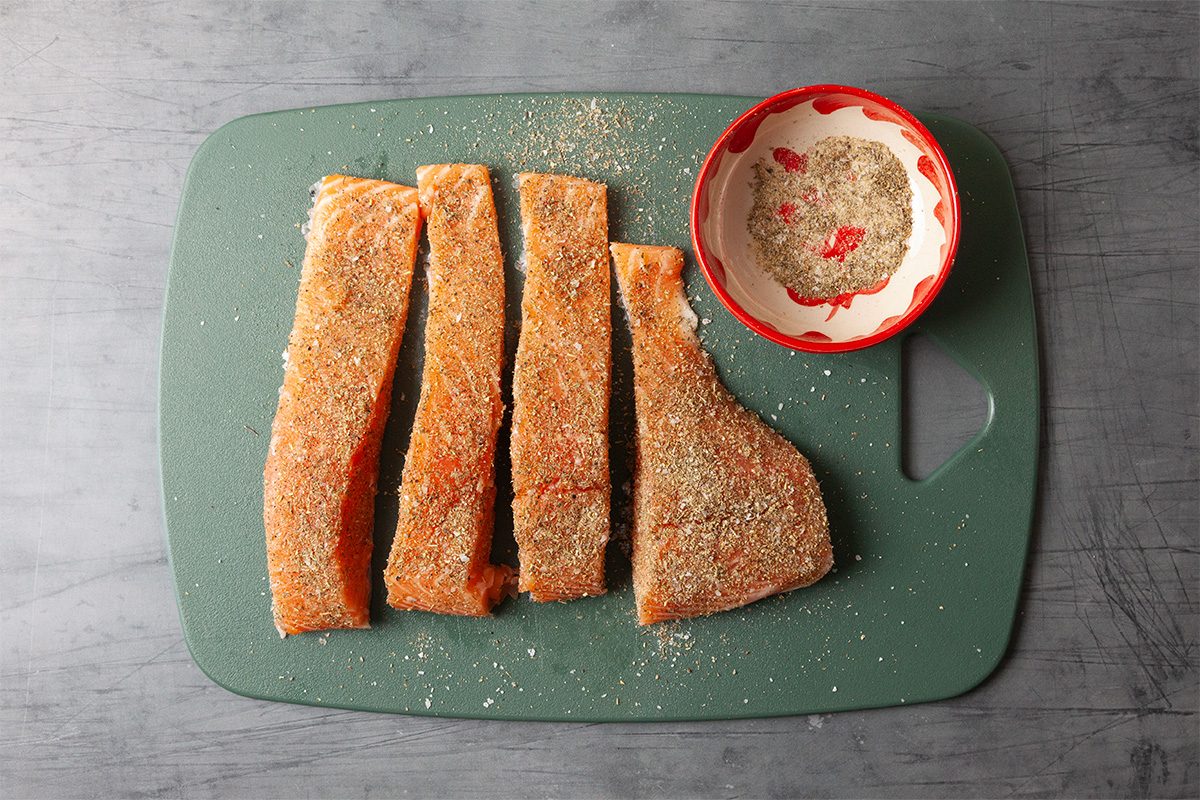 Overhead view of raw, seasoned salmon fillets arranged on a cutting board alongside a small bowl of spice mixture. The salmon is prepared with Italian seasoning, salt, pepper, and garlic powder, signaling the start of the cooking process.