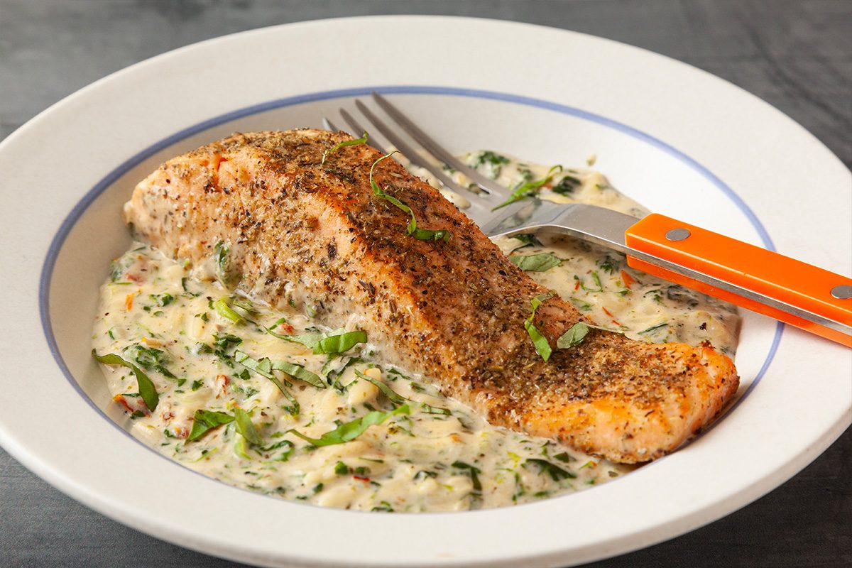 Tight overhead horizontal close-up of a single salmon fillet served over orzo pasta and creamy spinach sauce, garnished with fresh basil.