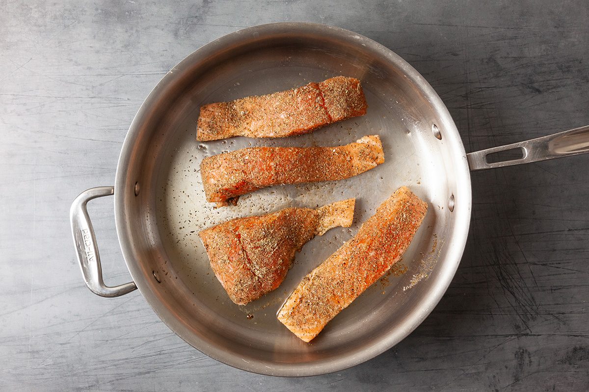 Overhead image of salmon fillets searing in a skillet with olive oil. The fish develops a golden crust as it cooks, showing the initial stovetop cooking stage.