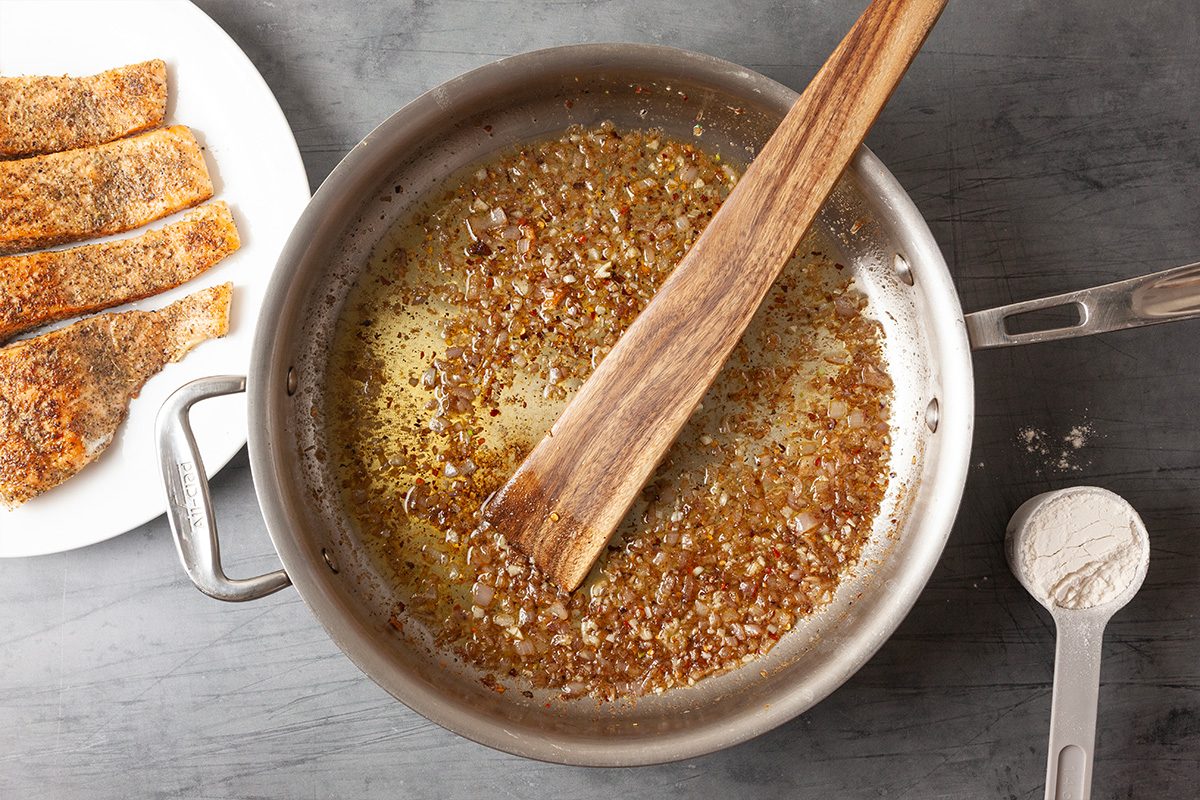 Overhead view of butter melting in a skillet with minced shallot and garlic. A wooden spoon stirs the aromatics as the sauce base begins to develop.