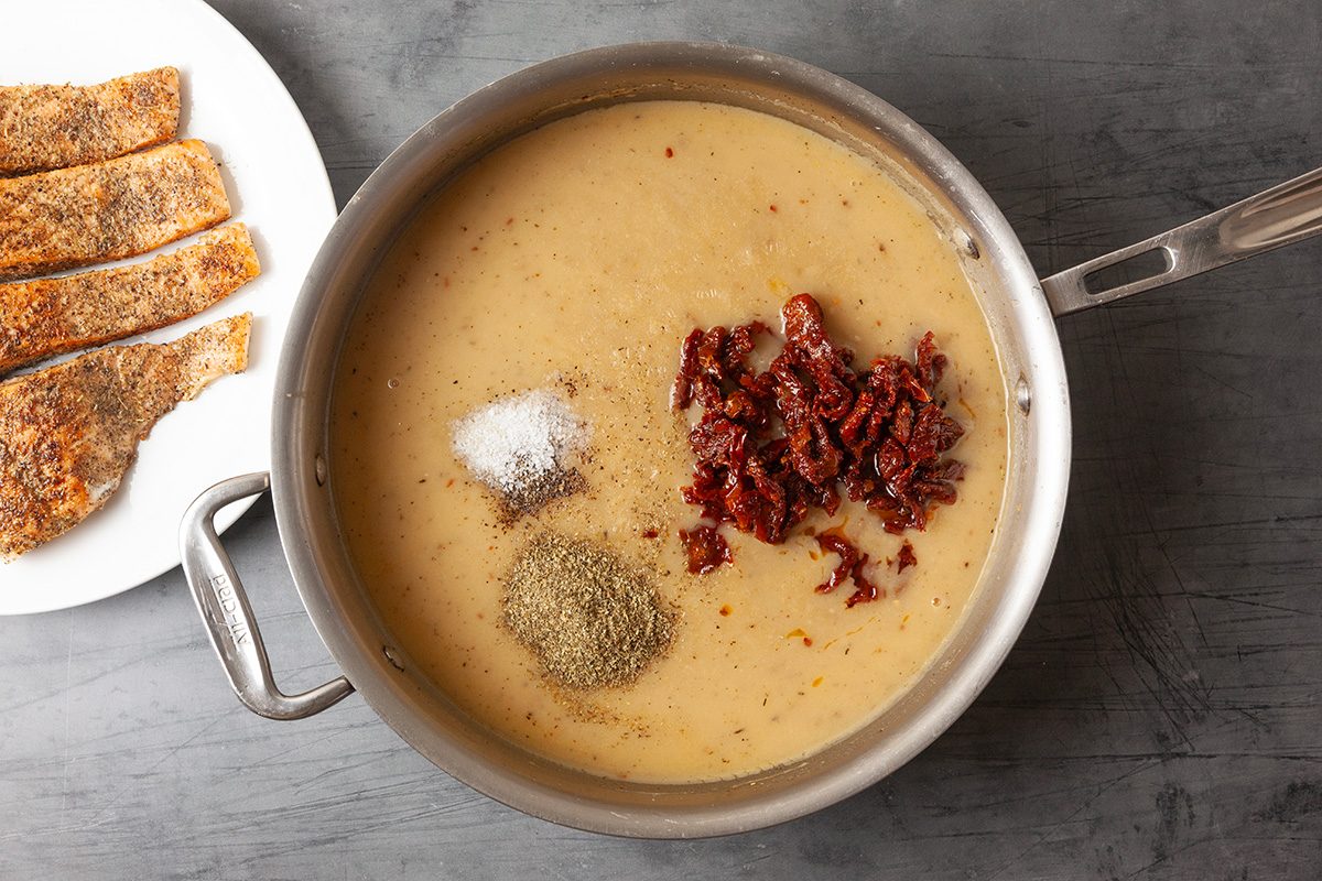 Overhead image of a creamy sauce base in a skillet with seasonings and crushed red pepper flakes added. The mixture simmers as flavor components are incorporated.