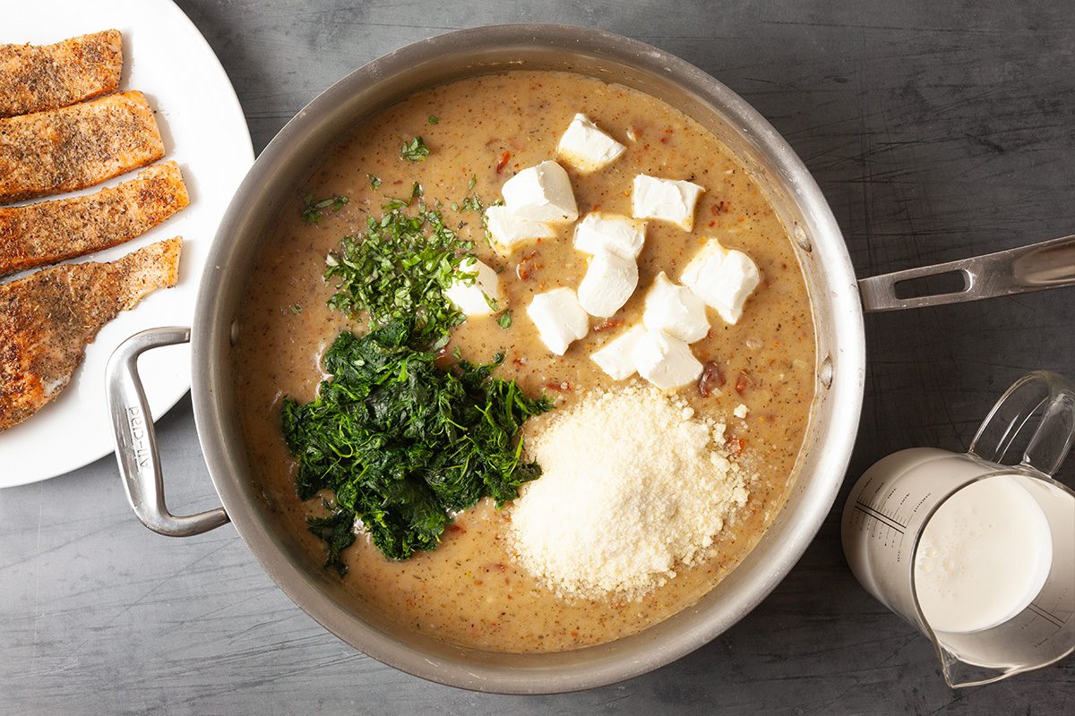 Overhead view of flour, cream cheese, spinach, Parmesan cheese, and fresh basil added to the sauce. Ingredients are layered before being stirred into a rich, creamy mixture.