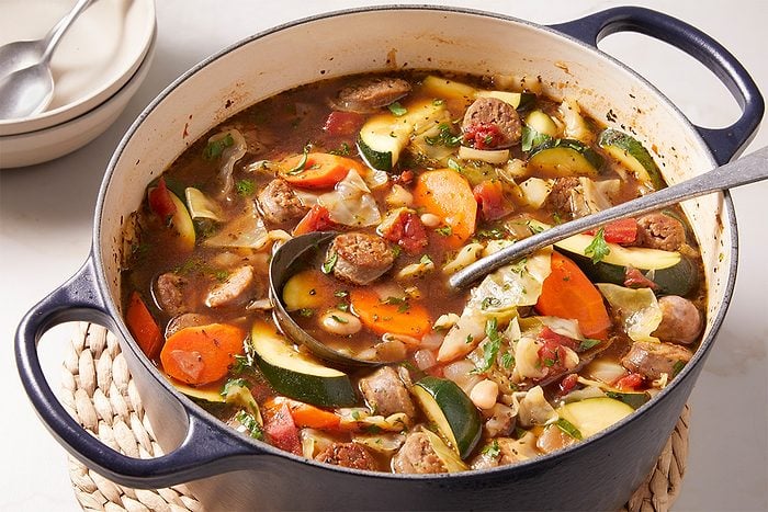 Close-up overhead shot of Minestrone Soup in a pot with a ladle resting inside, highlighting sliced carrots, zucchini, beans, and herbs floating in the hearty broth.